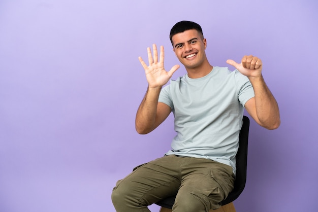 Jeune homme assis sur une chaise sur un mur isolé comptant six avec les doigts