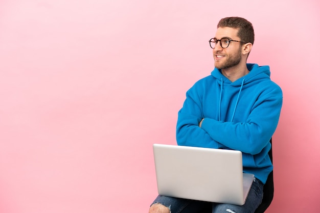 Jeune homme assis sur une chaise avec un ordinateur portable avec les bras croisés et heureux