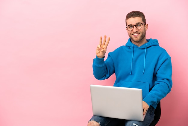 Jeune homme assis sur une chaise avec un ordinateur portable heureux et en comptant trois avec les doigts