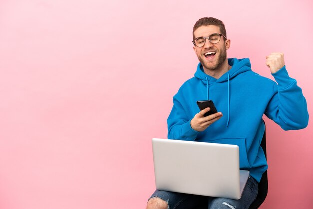 Jeune homme assis sur une chaise avec ordinateur portable avec téléphone en position de victoire