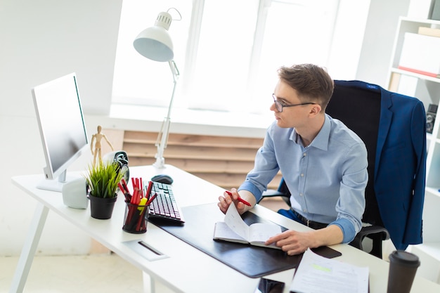 Un jeune homme assis dans le bureau au bureau de l'ordinateur tient un stylo à la main et regarde l'écran.