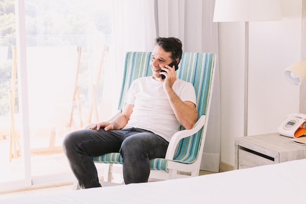 Jeune homme assis dans une chaise berçante dans sa chambre parlant au téléphone portable et souriant avec la lumière traversant la fenêtre Concept de connexion vacances-travail et smartphone