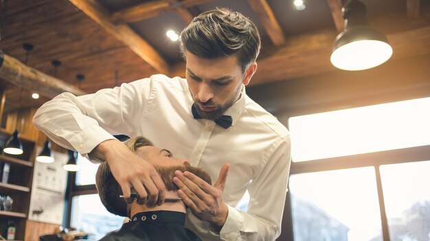 Jeune homme assis dans un salon de coiffure pendant que le coiffeur travaille avec un rasoir