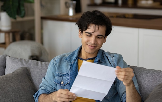 Jeune Homme Assis Lisant De Bonnes Nouvelles Dans Une Notification Papier Une Lettre D'embauche Pour Un Nouvel Emploi Ou Un Excellent Résultat D'examen Sur Le Canapé à La Maison