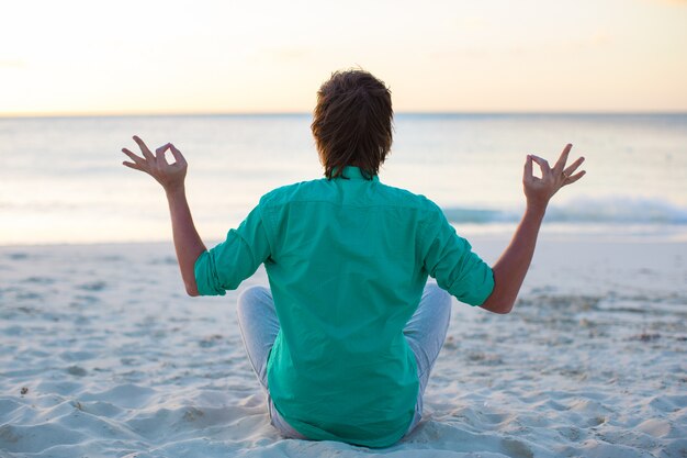 Jeune homme assis en position du lotus sur la plage de sable blanc