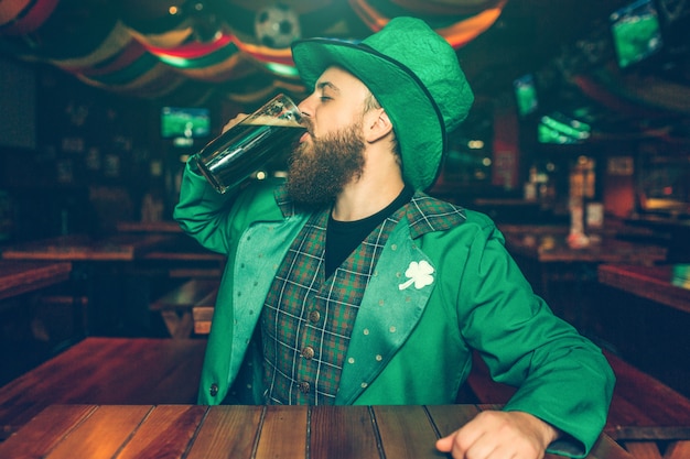 Un jeune homme assis seul à table dans un pub. Il boit de la bière brune dans une tasse. Guy porte le costume de St. Patrick.