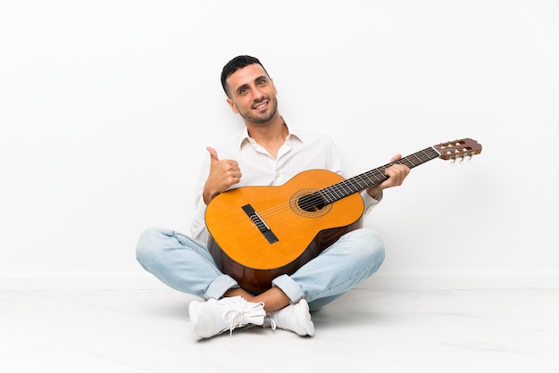 Jeune homme assis sur le sol avec une guitare avec le pouce levé parce qu'il s'est passé quelque chose de bien