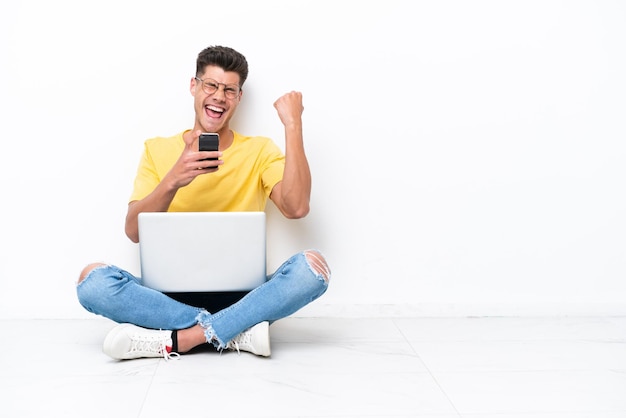 Jeune homme assis sur le sol isolé sur fond blanc avec téléphone en position de victoire