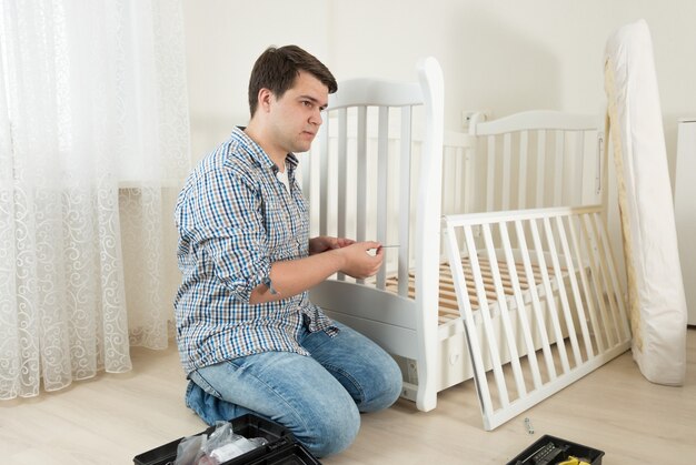 Jeune homme assis sur le sol et réparant le lit de l'enfant en pépinière