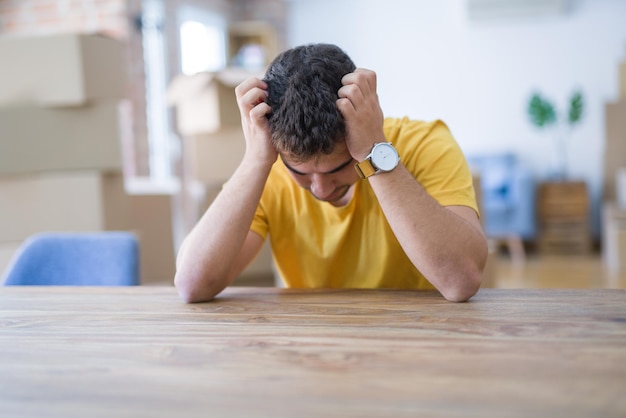 Jeune homme assis sur la table avec des boîtes en carton derrière lui déménageant dans une nouvelle maison souffrant de maux de tête désespérés et stressés à cause de la douleur et de la migraine Mains sur la tête