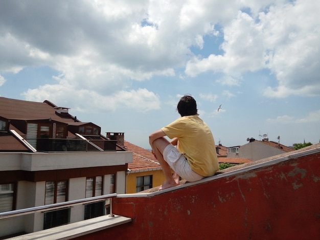 Photo jeune homme assis sur le toit de la maison, profitant de la vue sur les mouettes de la vieille ville volant dans un ciel d'été nuageux