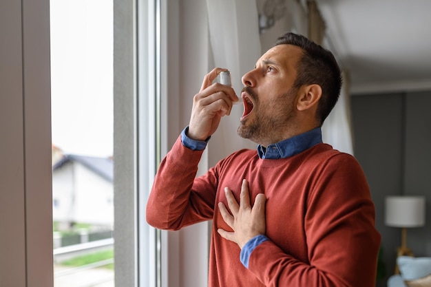 Jeune homme asthmatique touchant la poitrine dans la douleur et inhalant par la pompe à asthme par la fenêtre à la maison