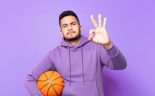 Jeune homme athlète hispanique expression heureuse et tenant un ballon de basket