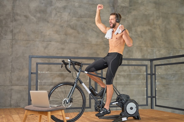 Jeune homme athlétique levant la main et criant de joie en faisant du vélo d'exercice