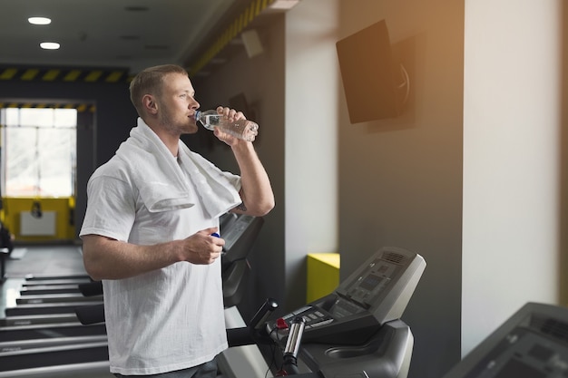 Jeune homme athlétique se reposant après l'entraînement, buvant de l'eau au gymnase, debout sur un tapis roulant, espace de copie. Concept de musculation