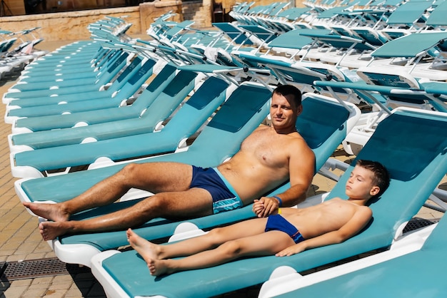 Un jeune homme athlétique et son fils sourient joyeusement et prennent un bain de soleil sur une chaise longue par une journée ensoleillée à l'hôtel Joyeuses vacances en famille dans un hôtel de la station Vacances d'été et tourisme