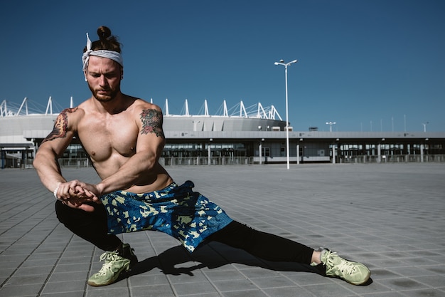 Un jeune homme athlétique avec un torse nu avec un bandeau vêtu de leggings noirs et d'un short bleu fait des étirements sur des dalles de pavage sur le fond du stade par une chaude journée ensoleillée.