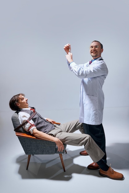 Photo jeune homme attaché à une chaise avec une ceinture en visite chez le dentiste médecin médecin tenant une seringue avec un fou fou