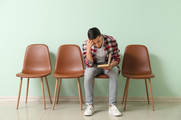 Jeune homme en attente avec un livre assis sur une chaise près du mur de couleur