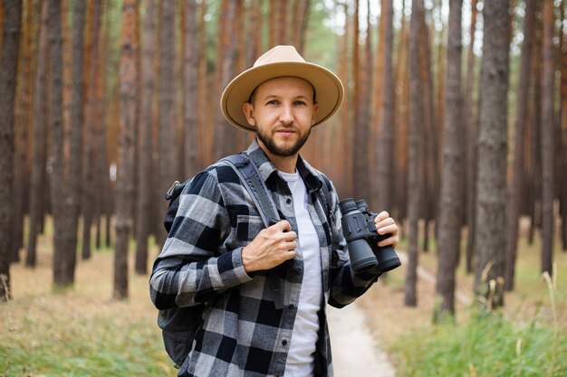 Jeune homme au chapeau avec un sac à dos et des jumelles dans une forêt de pins. Randonnée en montagne ou en forêt.