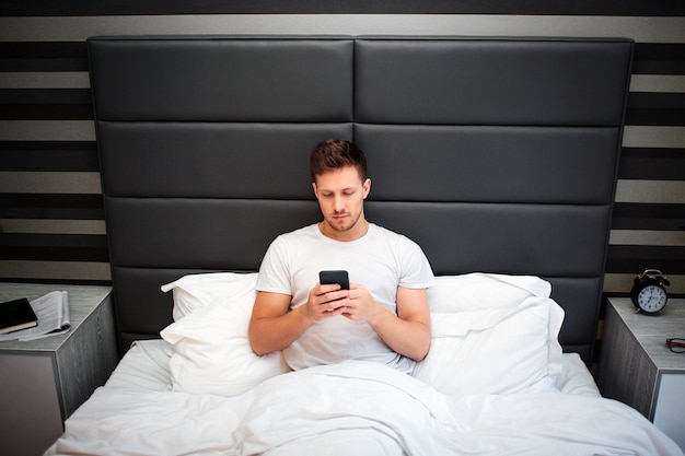 Jeune homme au lit ce matin. Il tient le téléphone. Guy regarde-le. Homme sérieux et concentré. Heure du coucher.