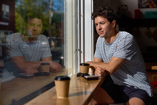 Jeune homme aux cheveux bouclés assis près de la fenêtre dans un café par une journée ensoleillée, verres en papier avec cappuccino, style de vie