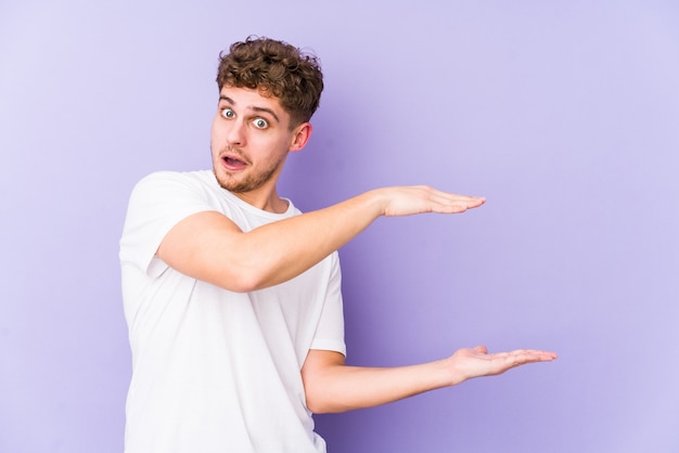 Jeune homme aux cheveux bouclés blonds choqué et étonné tenant quelque chose entre les mains