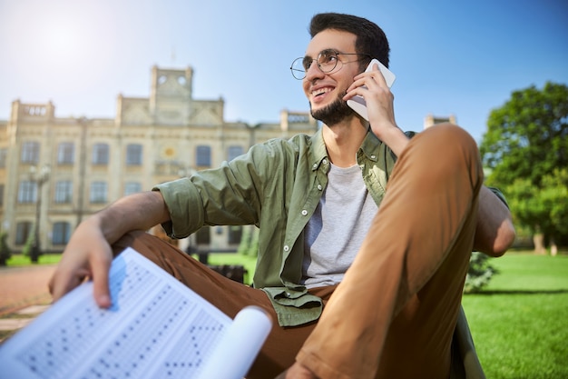 Jeune homme aux cheveux courts barbu attrayant joyeux dans des lunettes rondes parlant sur son téléphone portable à l'extérieur