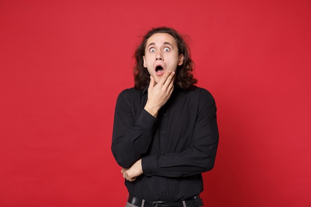 Jeune homme aux cheveux longs bouclés triste en chemise noire debout posant isolé sur fond de mur rouge portrait en studio. Concept de mode de vie des émotions sincères des gens. Maquette de l'espace de copie. Dépression de crise de problème