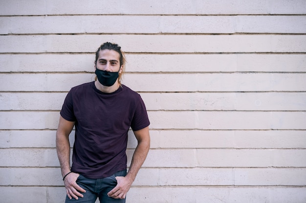 Jeune homme aux cheveux longs debout sur un mur tout en portant un masque noir contre la pandémie de coronavirus