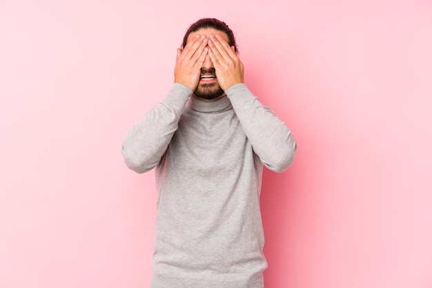 Jeune homme aux cheveux longs isolé sur un rose couvre les yeux avec les mains, sourit largement en attente d'une surprise.