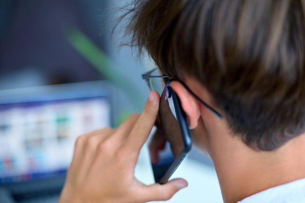 Jeune homme aux cheveux noirs et lunettes parlant au téléphone et regardant un ordinateur portable