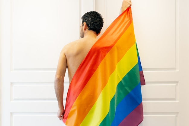 Photo jeune homme aux cheveux noirs par derrière avec dos nu recouvert de lgbtqi + drapeau arc-en-ciel au centre de l'image avec portes fermées blanches