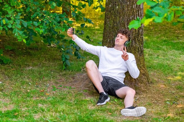 Jeune homme aux cheveux roux dans les écouteurs assis appuyé sur un arbre en prenant un selfie