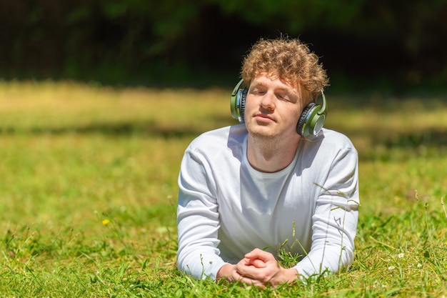 Jeune homme aux cheveux roux dans les écouteurs se trouve sur l'herbe verte sur une journée ensoleillée