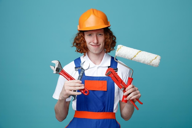 Jeune homme avide de constructeur en uniforme tenant des outils de construction isolés sur fond bleu