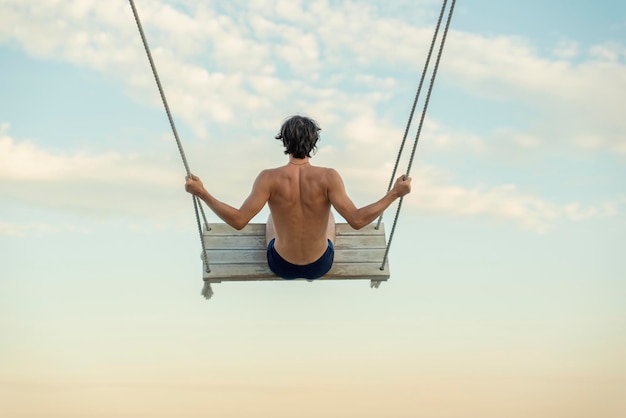 Jeune homme sur une balançoire haut dans le ciel sur fond de nuages Guy se balançant vue arrière Lumière douce du coucher du soleil