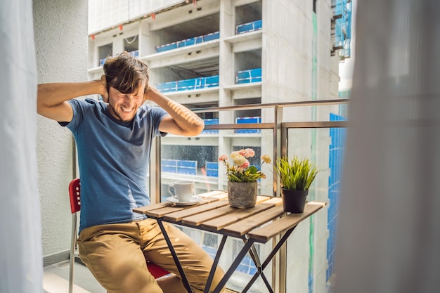 Jeune homme sur le balcon agacé par les travaux de construction à l'extérieur Concept de bruit Pollution de l'air par la poussière de construction