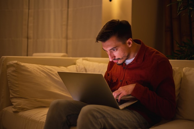 Un jeune homme avec une barbe en chemise rouge travaille avec un ordinateur portable et s'assoit sur le canapé le soir à la maison.