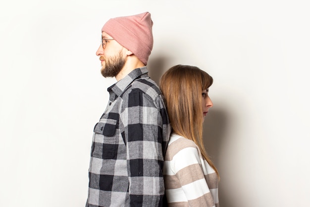 jeune homme avec une barbe dans un chapeau et une chemise à carreaux et une fille dans un pull se tiennent dos à dos sur un fond clair isolé.