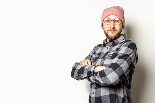 jeune homme avec une barbe dans un chapeau, une chemise à carreaux et des lunettes avec les bras croisés sur sa poitrine sur un blanc isolé.