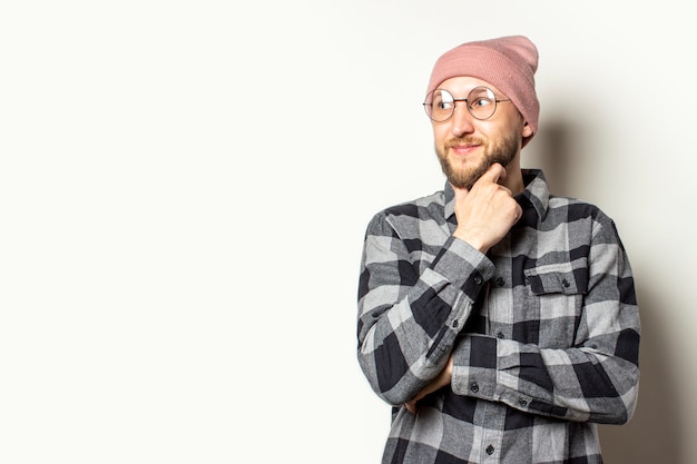 jeune homme avec une barbe dans un chapeau, une chemise à carreaux et des lunettes regarde ailleurs avec un visage gêné sur un blanc isolé.