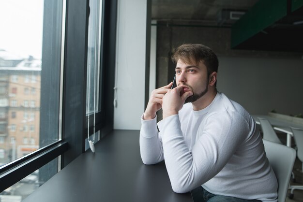 Un jeune homme avec une barbe est assis dans un bureau moderne à la table près de la fenêtre et parle au téléphone.