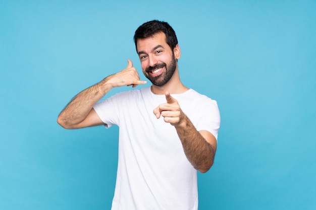 Jeune homme à la barbe sur fond bleu isolé, faisant un geste de téléphone et avant de pointage