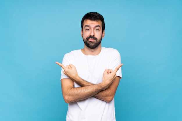 Jeune homme à la barbe sur mur bleu isolé, pointant vers les côtés ayant des doutes