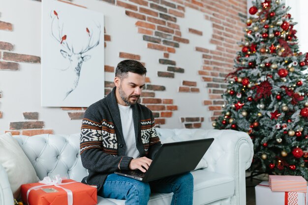 Jeune homme avec une barbe souriant et travaillant sur un ordinateur portable depuis la maison, assis sur le canapé pendant les vacances du Nouvel An