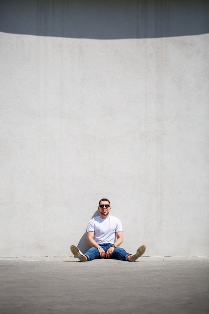 Un jeune homme avec une barbe en T-shirt est assis près d'un mur gris. Mec en lunettes de soleil