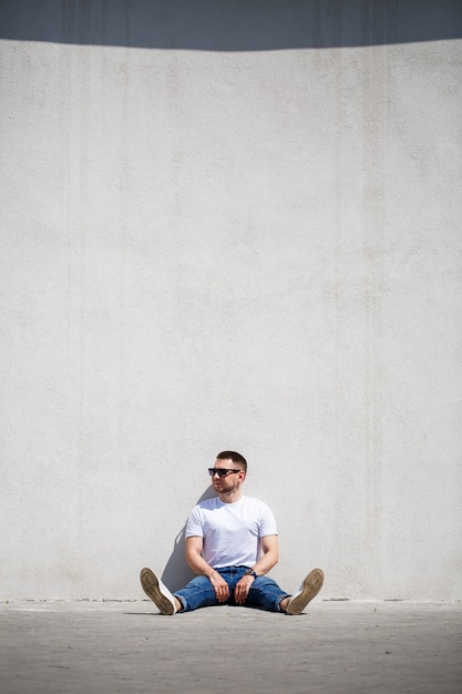 Un jeune homme avec une barbe en T-shirt est assis près d'un mur gris. Mec en lunettes de soleil