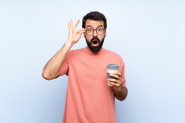 Jeune homme à la barbe tenant un café à emporter sur bleu isolé avec des lunettes et surpris