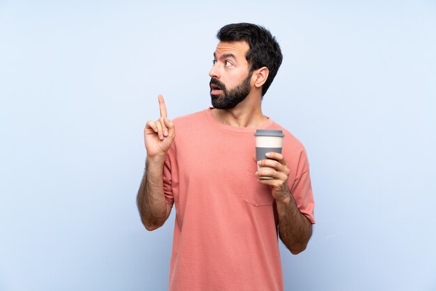 Jeune homme à la barbe tenant un café à emporter sur bleu isolé, pensant une idée pointant le doigt vers le haut